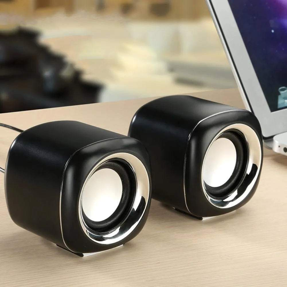 A pair of compact black wired desktop computer speakers with a 3.5mm audio cable on a wooden desk