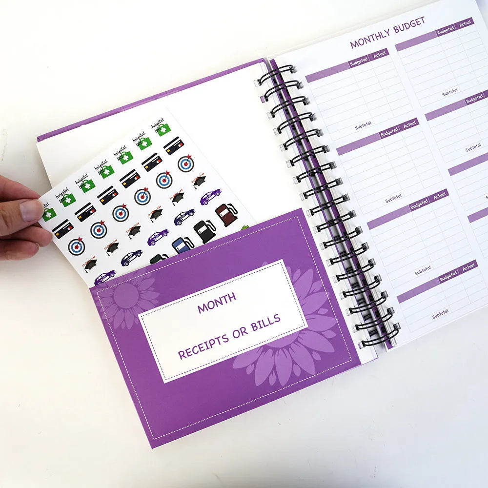 J12 Purple coil-bound monthly financial planning notebook with pen, stickers, and paper clips on a desk.