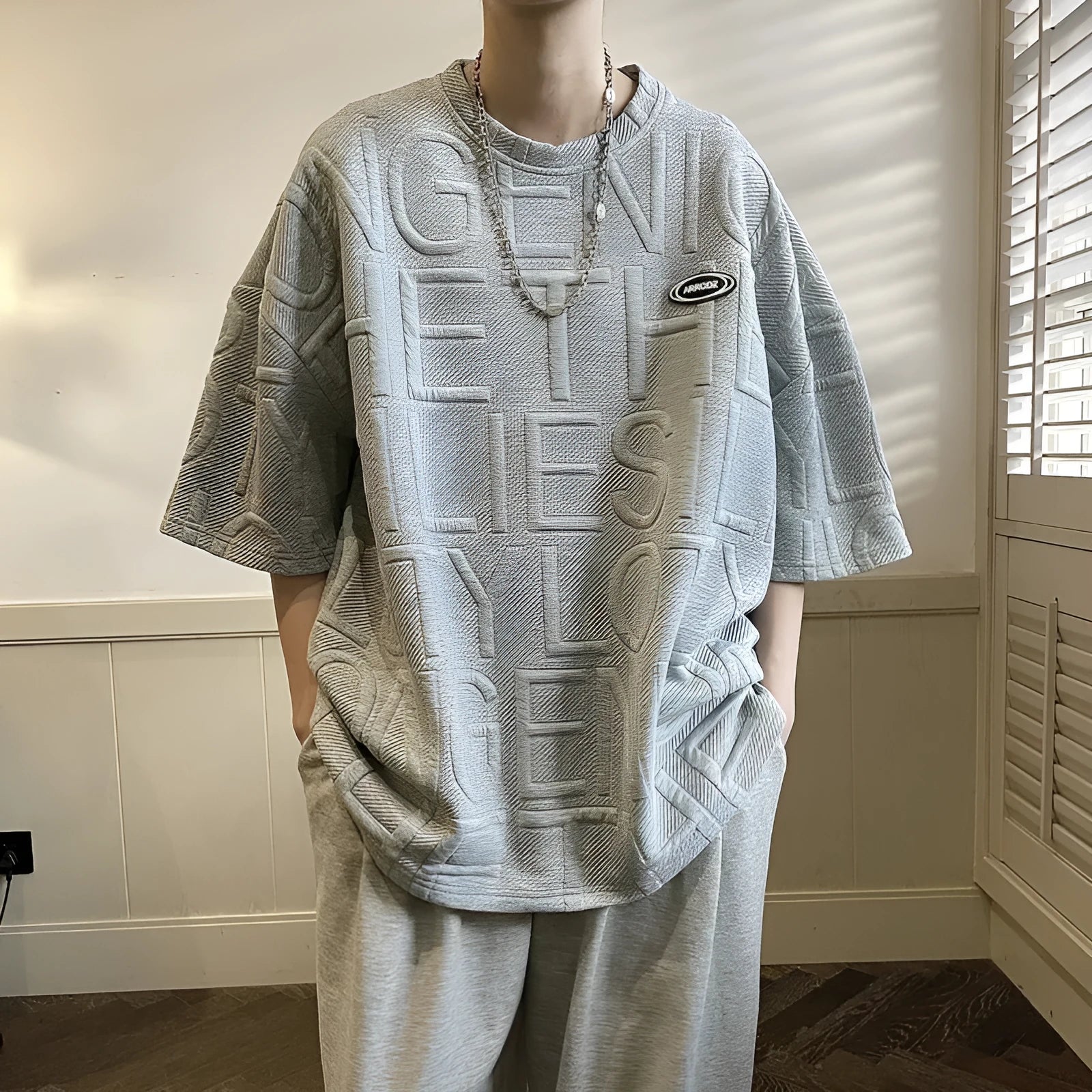 Men's grey short sleeve T-shirt with a raised 3D letter pattern, shown in a loose retro fit.